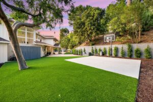 A beautiful home yard featuring vibrant trees and a basketball court bordered by realistic, green artificial turf