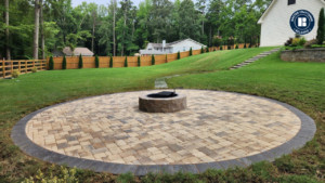 A wide backyard features a large, circular paver patio set into bright green grass. The patio is made of tan and gray interlocking pavers with a dark gray border ring, and a small round stone fire pit sits at the center. In the background, a wooden privacy fence lines the yard with tall trees beyond it, while a sloped lawn rises to the right with stone steps leading up toward a light-colored house and a short retaining wall. A “Master Craftsman Belgard” logo appears in the top-right corner.