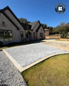 A modern brick house with black gabled roofs is seen from a low angle under a clear blue sky. In the foreground, a wide driveway features a gray herringbone-pattern paver section framed by light concrete edging, curving toward a smooth concrete area on the right. A strip of green grass borders the driveway, and trees and a neighboring building appear in the background. A circular “Master Craftsman Belgard” logo sits in the top-right corner.