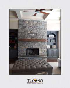 A living room centers on a tall, floor-to-ceiling stacked-stone fireplace in mixed gray tones. The stonework has a rugged, textured look, with a simple wood mantel running horizontally across the middle and a rectangular firebox opening near the bottom, finished with a stone hearth ledge. Built-in gray cabinets and shelves flank the fireplace on both sides, while a tufted ottoman sits in the foreground and a ceiling fan hangs above. The “TUCANO — Stone • Paver • Concrete” logo appears at the bottom of the image.